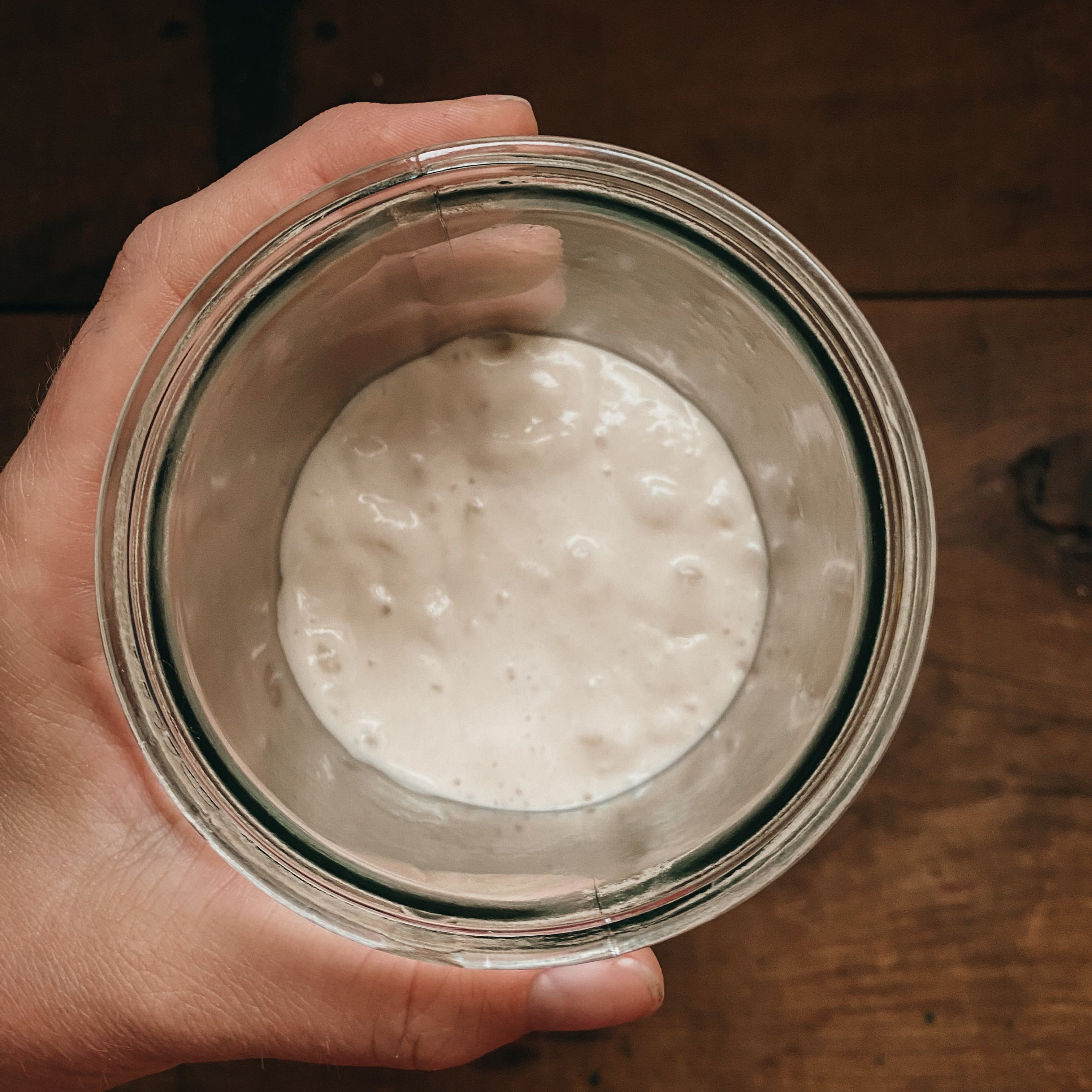 A hand holding a glass jar with a visible sourdough starter inside, on a wooden surface.