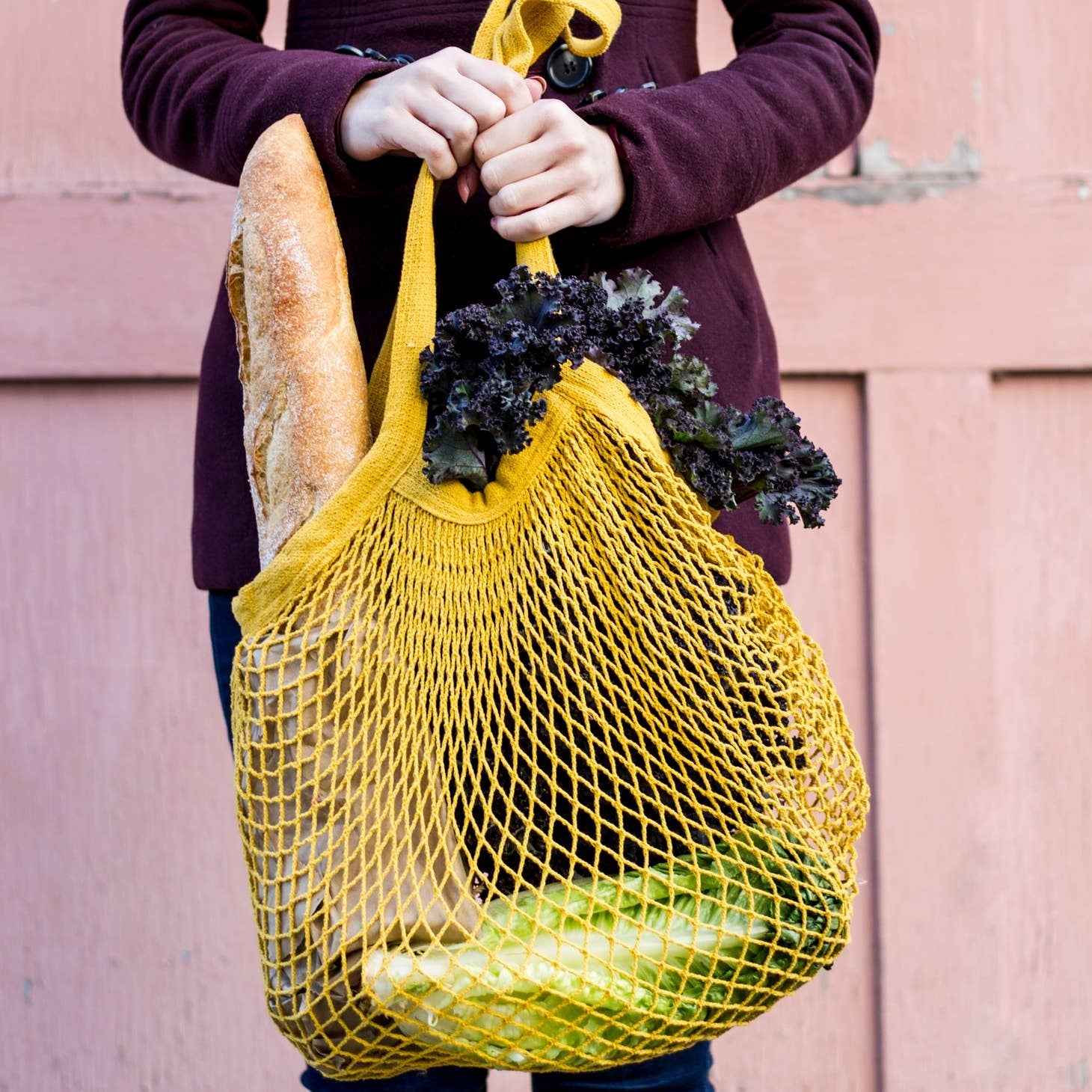 Mesh market bag with fresh vegetables, kale and a baguette, held by a person at a market