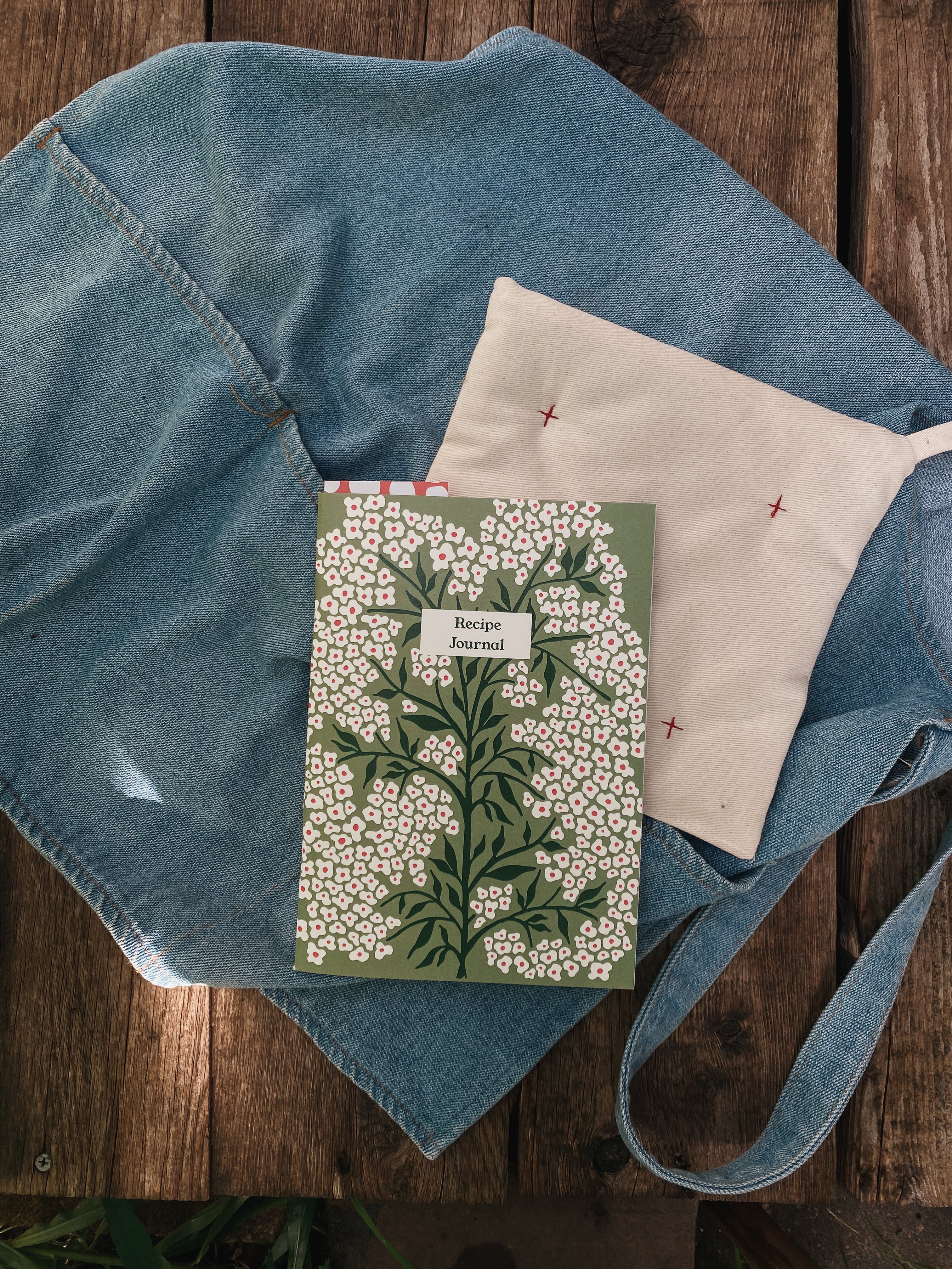 Picture of a forget-me-not recipe journal with an apron and a handmade potholder on a wooden background. 