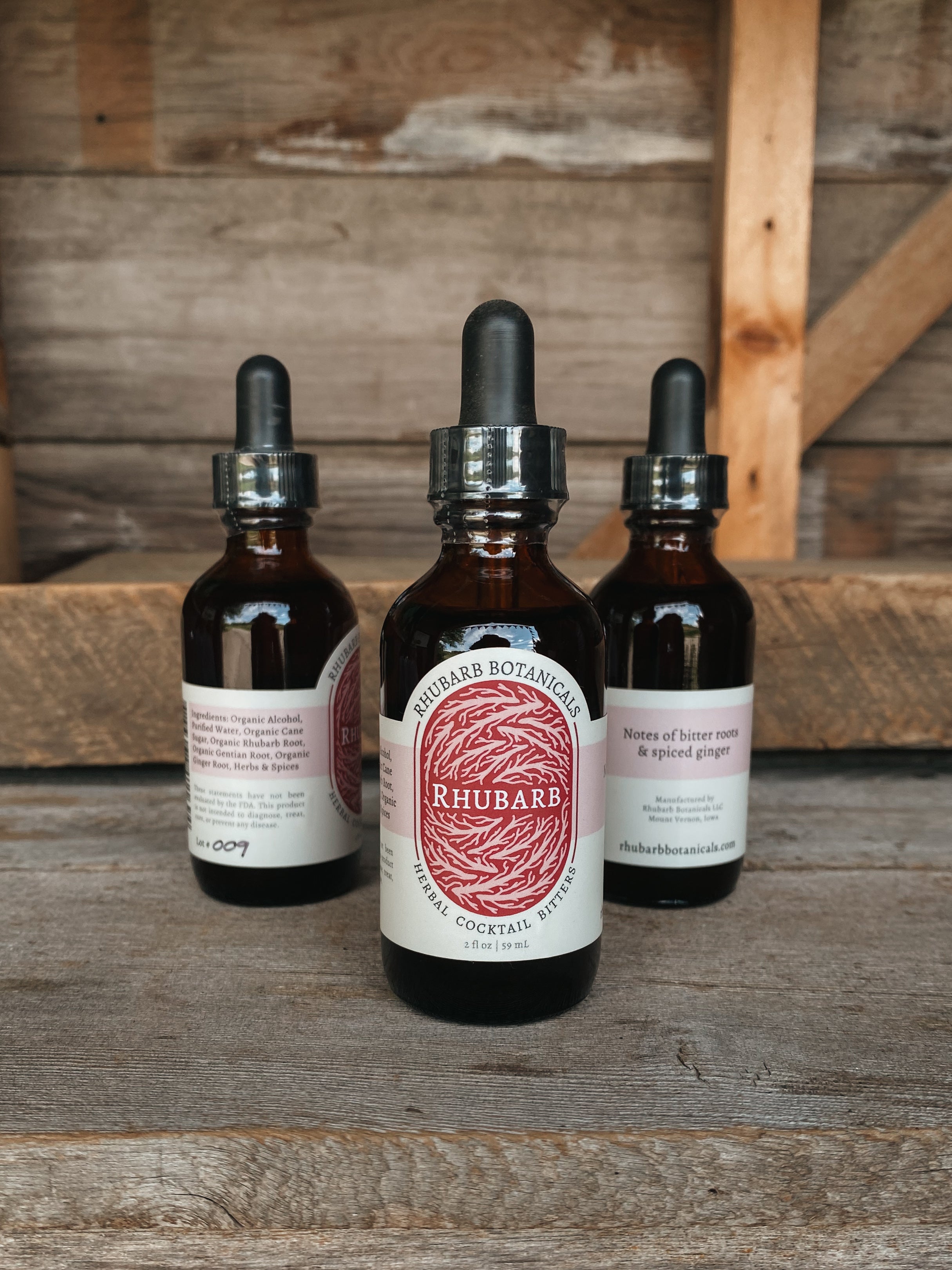 Three bottles of rhubarb cocktail bitters from Rhubarb Botanicals sit on a shelf with a rustic wooden background. 